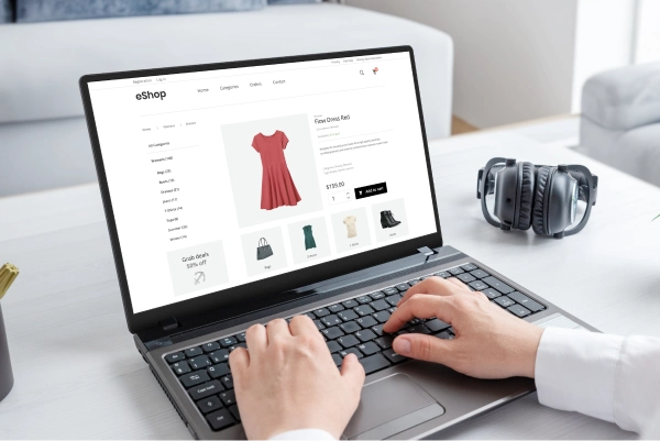 A person is shopping online using a laptop. The screen displays an e-commerce website featuring a red dress, with options to select size and add to cart. Nearby are headphones placed on the desk.