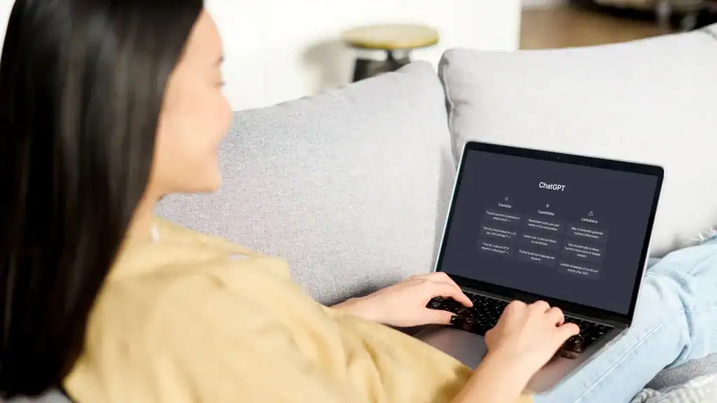 A woman sitting on a couch using a laptop, viewing the ChatGPT interface with example prompts, capabilities and limitations on the screen.