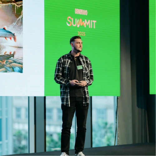 OMG Academy CEO speaking on stage at the General Summit 2025, standing in front of a large green event backdrop.