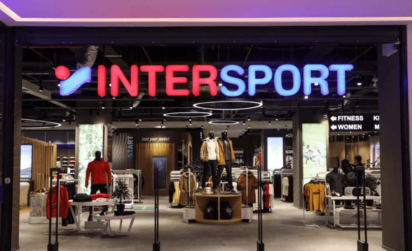 Intersport retail store front featuring the illuminated red and blue Intersport signage, mannequin displays with sportswear, and fitness and womens department wayfinding signs.
